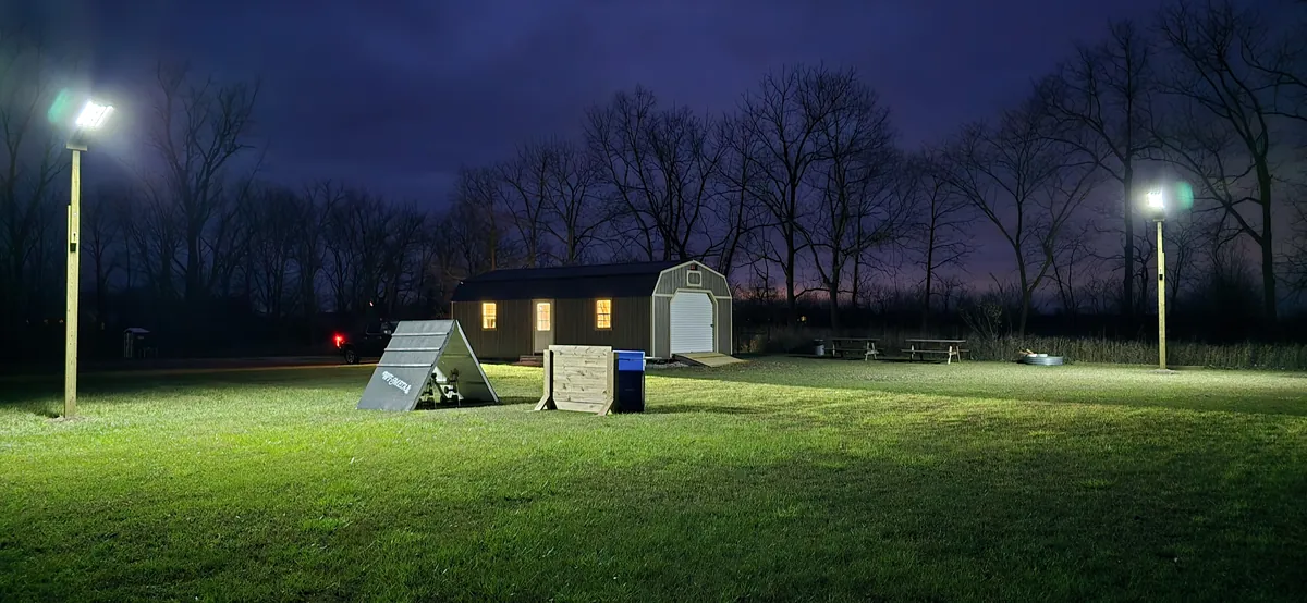 Barn-style shed lit at night in a large backyard with floodlights and dog agility equipment.