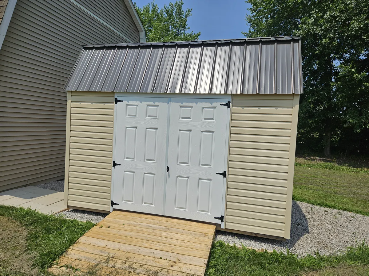 Beige shed with white double doors, a metal roof, and a wooden ramp beside a house.