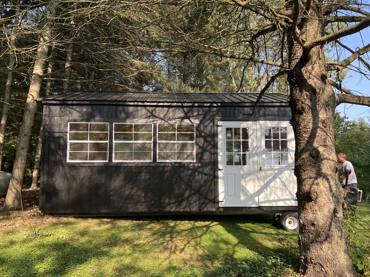 Black cabin-style shed with white double doors and large windows on a trailer under tall trees.