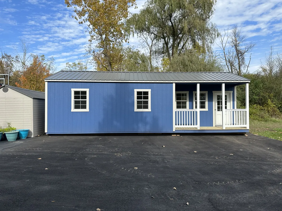 Blue cabin with a white porch, metal roof, and white-trimmed windows parked on an asphalt driveway.