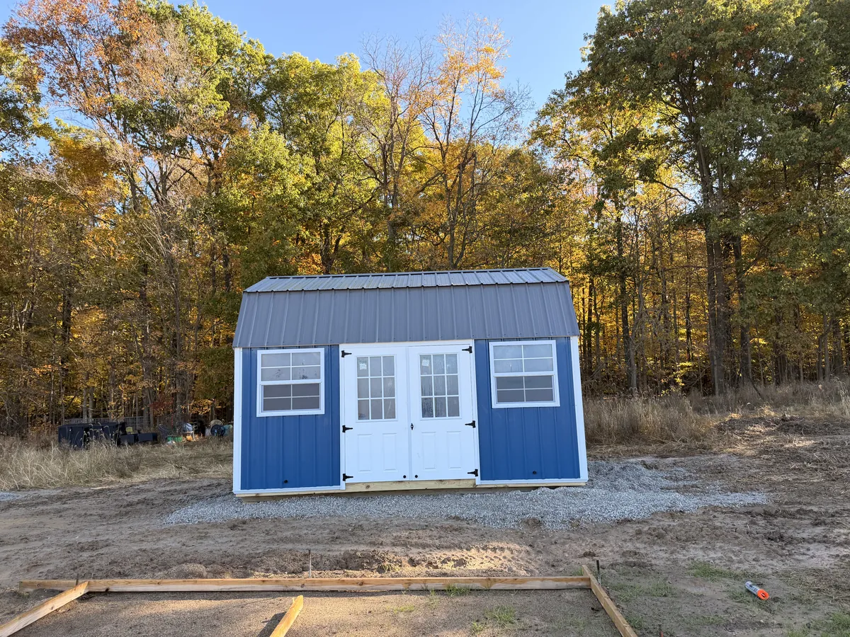 Blue lofted barn shed with white double doors and a gray metal roof on a freshly cleared lot with fall trees behind.