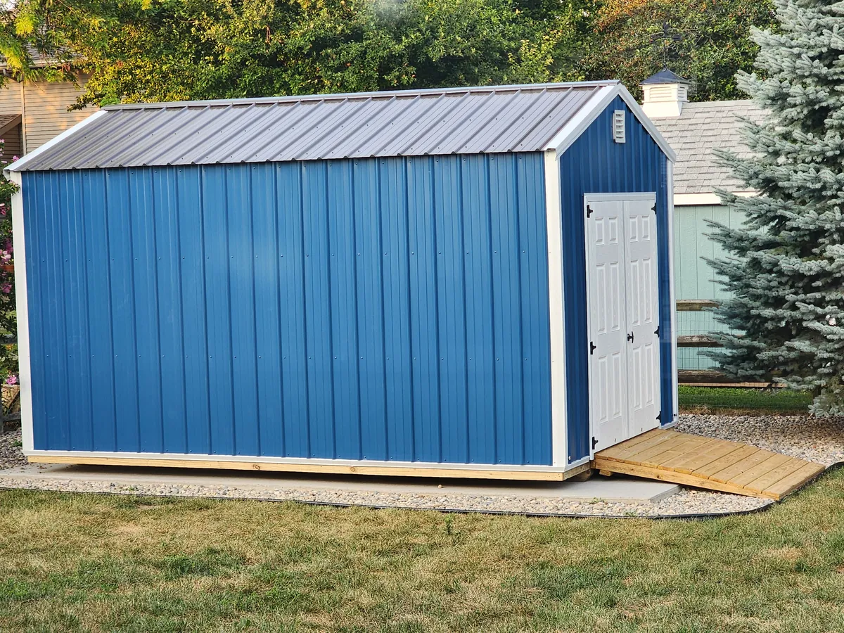 Blue metal-sided shed with a gray metal roof, white double doors, and a wooden ramp on a pad.