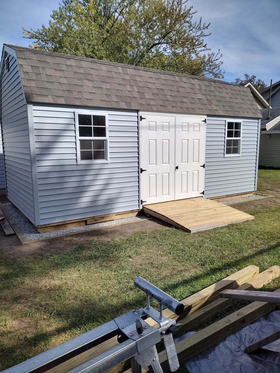 Light blue vinyl-sided shed with a shingled roof, white double doors, and a wooden ramp in a backyard.