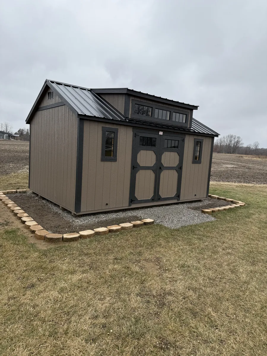 Brown shed with a black metal roof, transom dormer windows, and stone edging beside an open field.