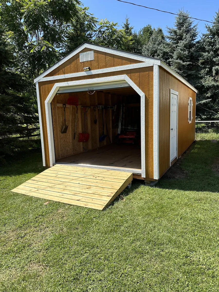 Brown garage shed with white trim, an open garage door, and a new wooden ramp on a lawn.