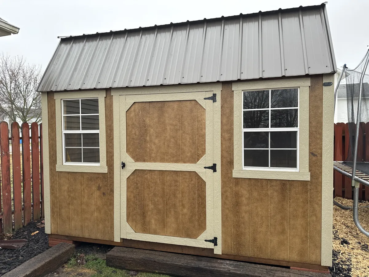 Wood-tone lofted barn shed with cream trim and a brown metal roof set between fences in a backyard.