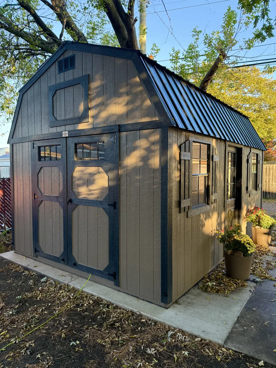 Brown lofted barn shed with black trim, black metal roof, and shuttered windows on a concrete pad.