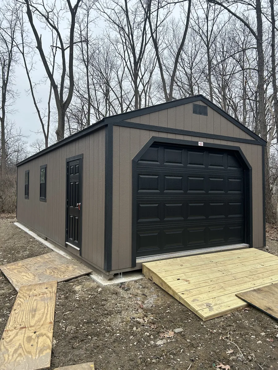 Brown portable garage with a black overhead door and wooden ramp on a wooded lot.