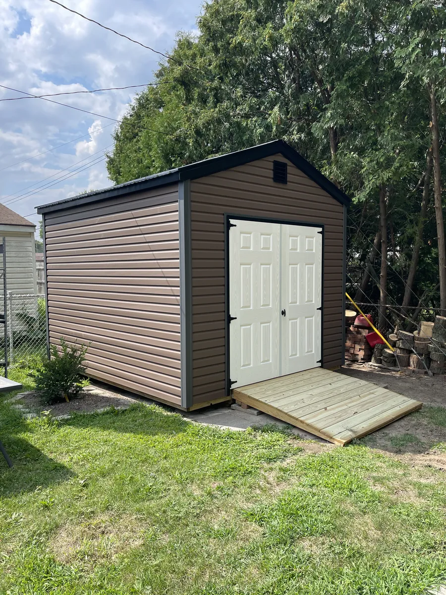 Brown utility shed with white double doors and a wooden ramp in a backyard near trees.