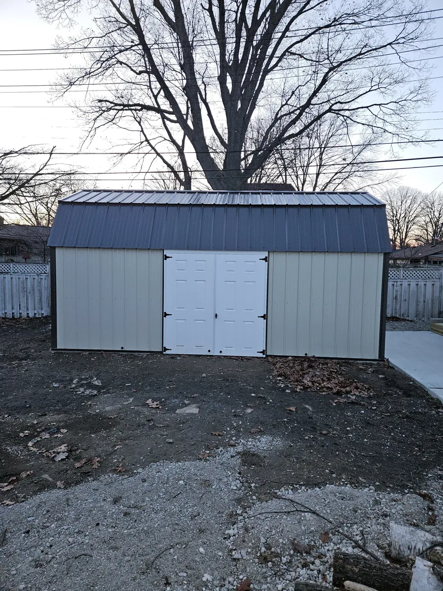 Cream lofted barn shed with a black metal roof and white double doors on a graded dirt yard.