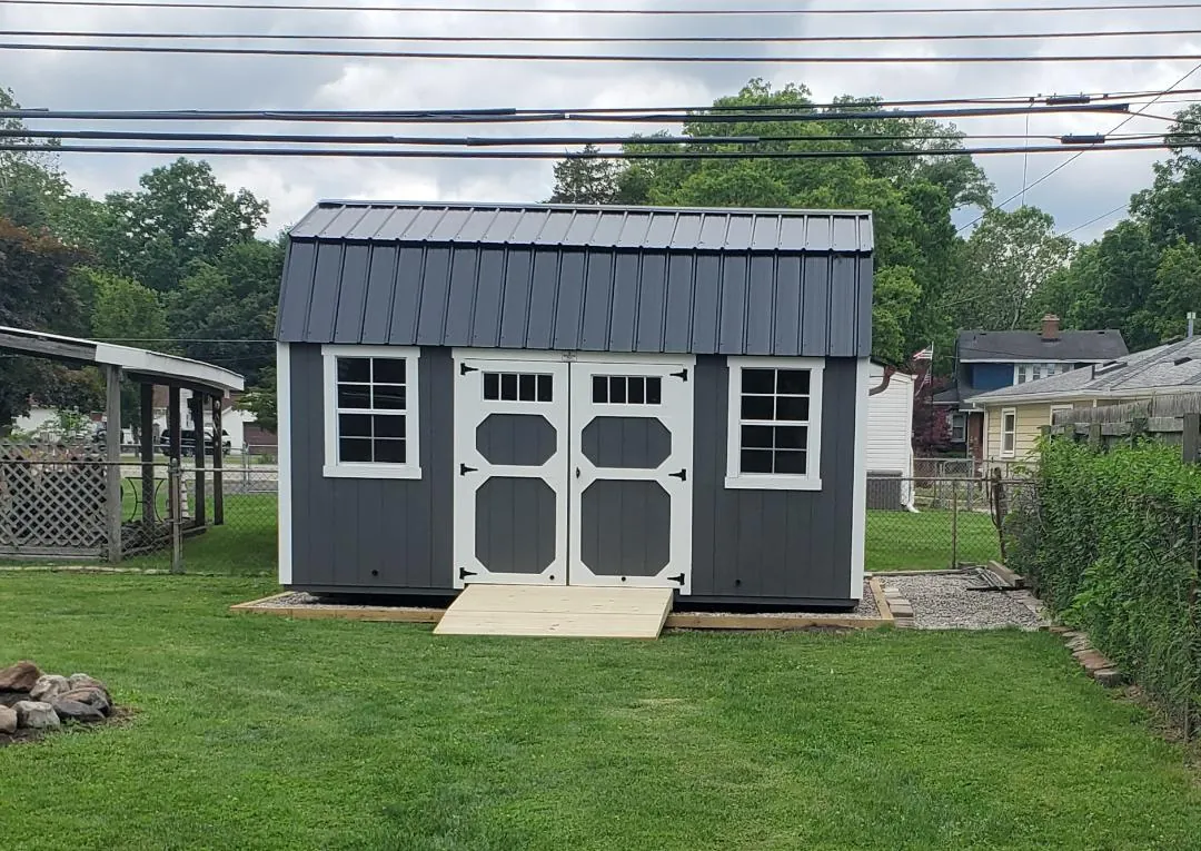 Dark gray lofted barn shed with white trim, double doors, and a ramp on a backyard lawn.