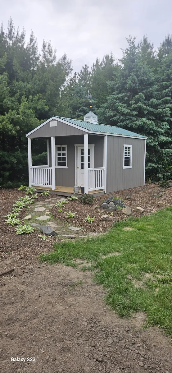 Gray cabin-style shed with front porch, white railings, and cupola set among evergreen trees.