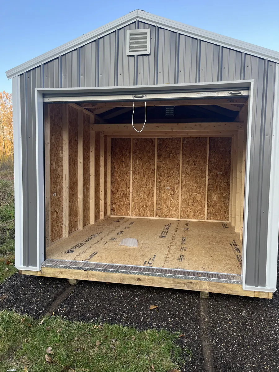 Gray metal garage shed with the roll-up door open, showing an unfinished interior with exposed framing.