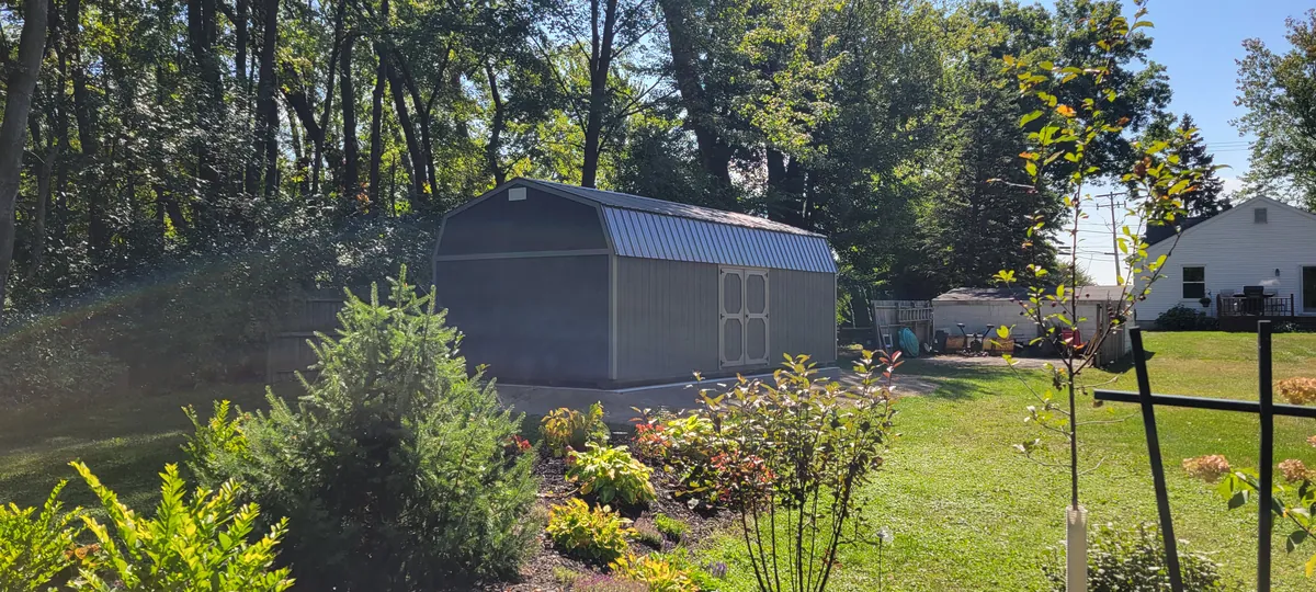 Gray lofted barn shed with a metal roof behind a flower garden in a wooded yard.