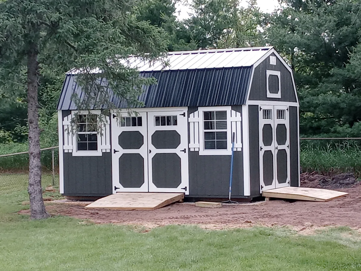 Dark gray lofted barn shed with white trim, two sets of doors, and wooden ramps in a backyard.