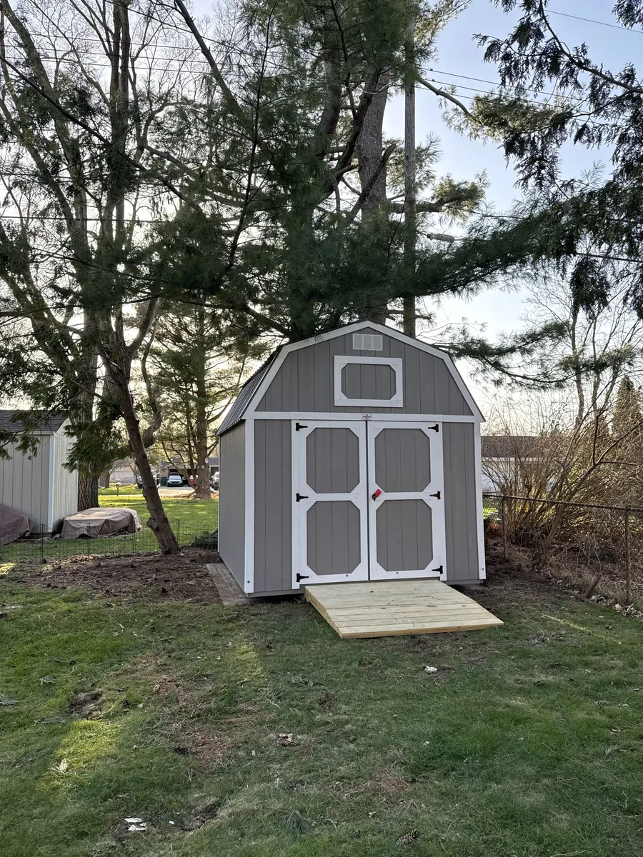 Gray lofted barn shed with white trim and a wooden ramp beneath tall trees.