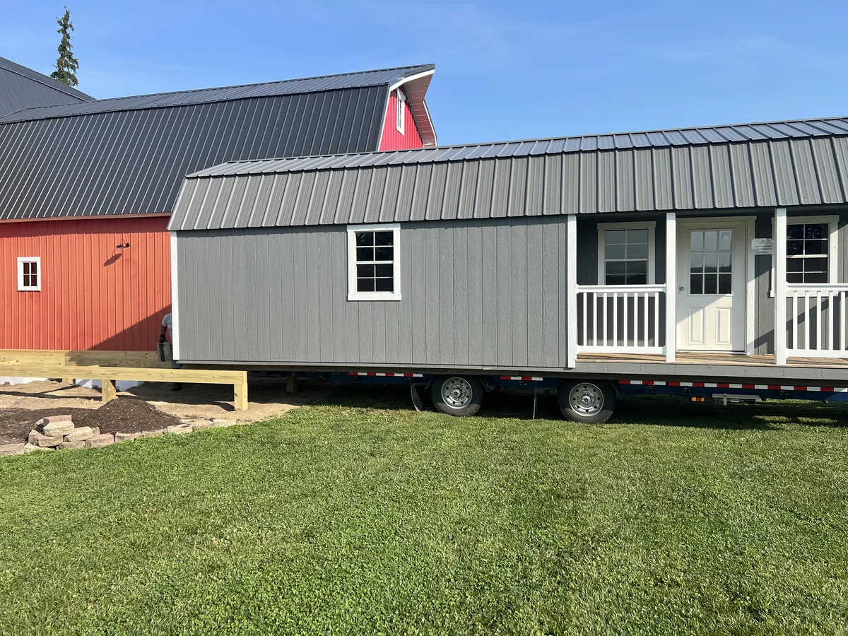 Gray lofted barn cabin with a porch on a delivery trailer in front of a red barn.