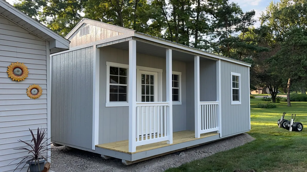 Gray cabin-style shed with a covered front porch and white railings next to a house in a backyard.
