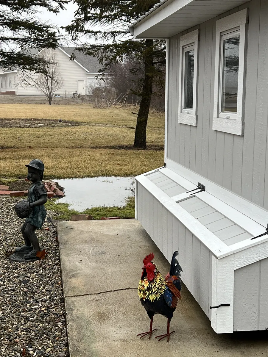 Side of a gray shed with an attached storage box, next to garden statues and a metal rooster.