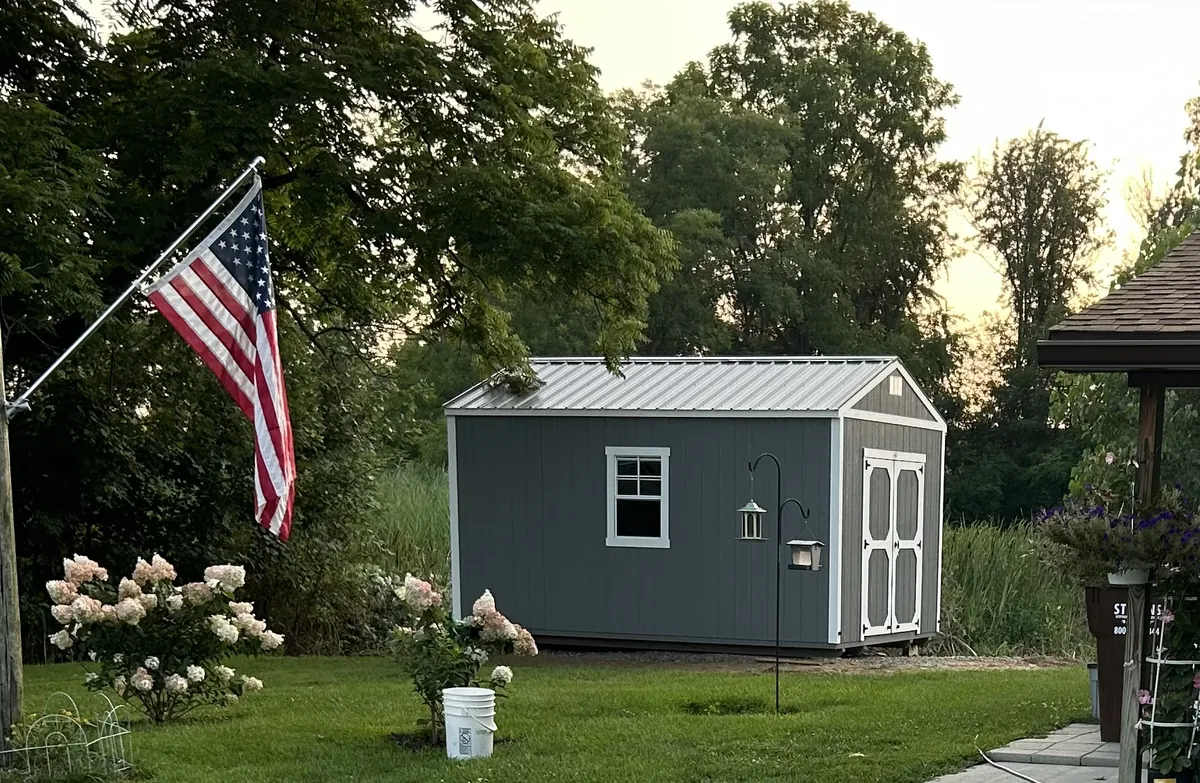Gray shed with white trim and a metal roof in a backyard with an American flag and flowers.