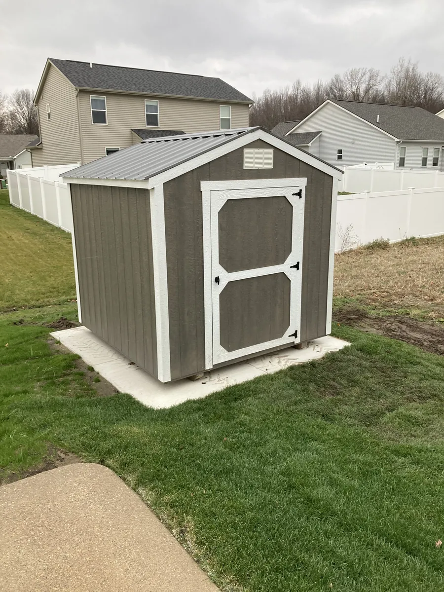Gray utility shed with white trim and a metal roof on a concrete pad in a subdivision backyard.