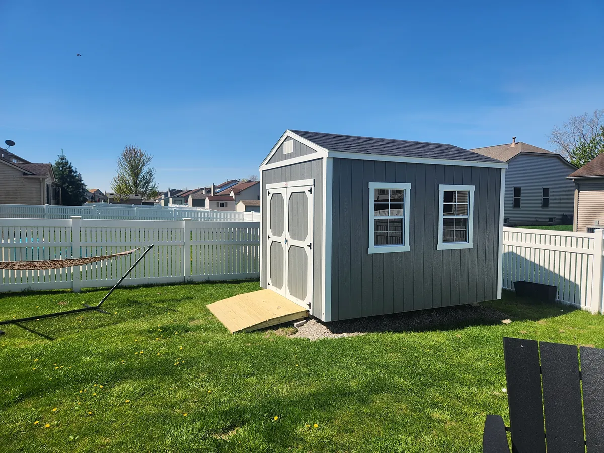Gray utility shed with white trim, two windows, and a wooden ramp in a fenced backyard.
