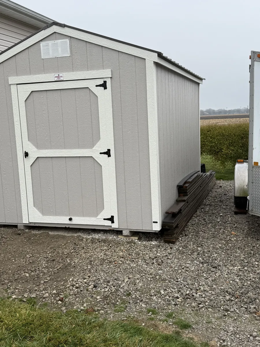 Gray utility shed with a white trim door on a gravel pad beside a farm field.
