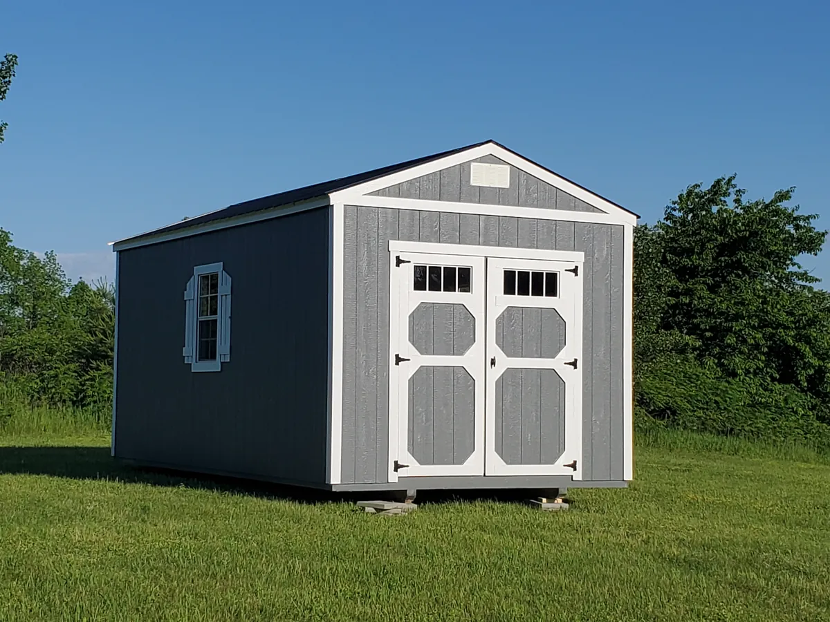 Gray utility shed with white double doors and transom windows on a mowed lawn beside an open field.