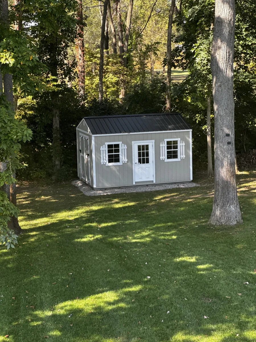 Gray utility shed with white door, shuttered windows, and black metal roof on a shaded backyard lawn.