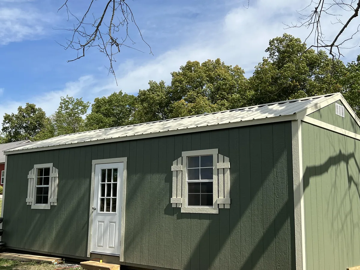Green cottage-style shed with white trim windows and a single entry door, viewed from the side under shade trees.