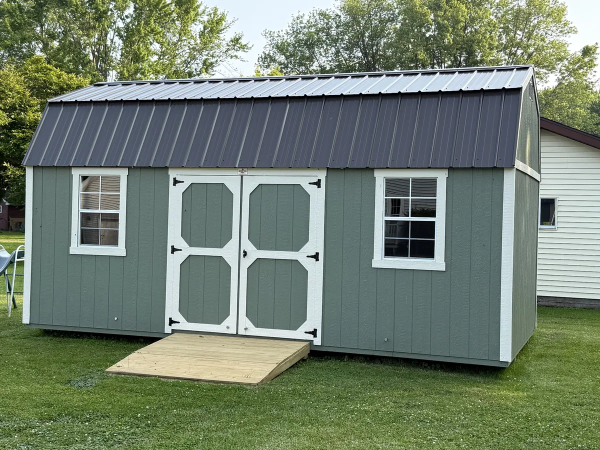 Sage green lofted barn shed with white trim, a black metal roof, and a wooden ramp on a lawn.
