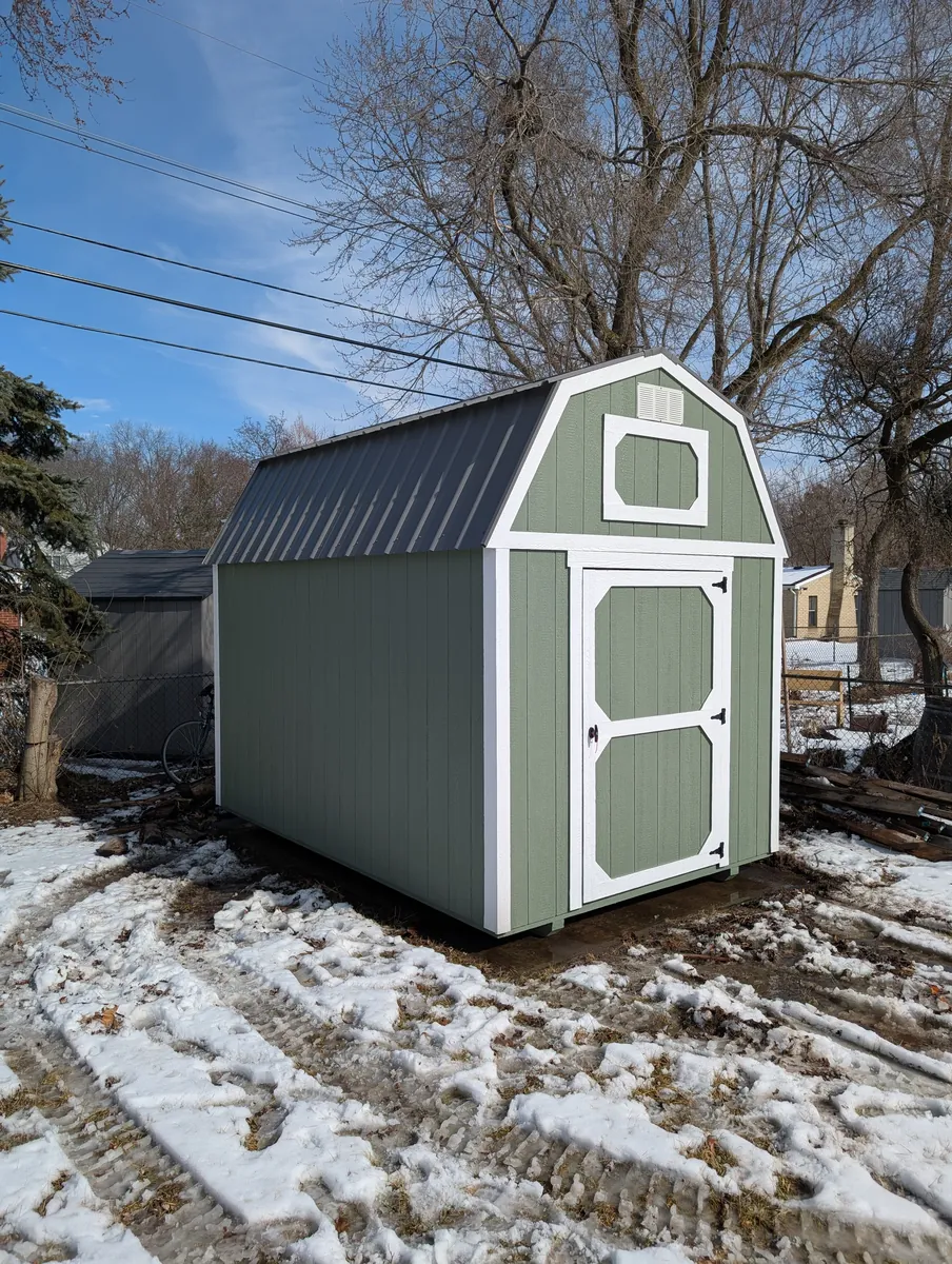 Green lofted barn shed with white trim and single door on a muddy lot with patches of snow.