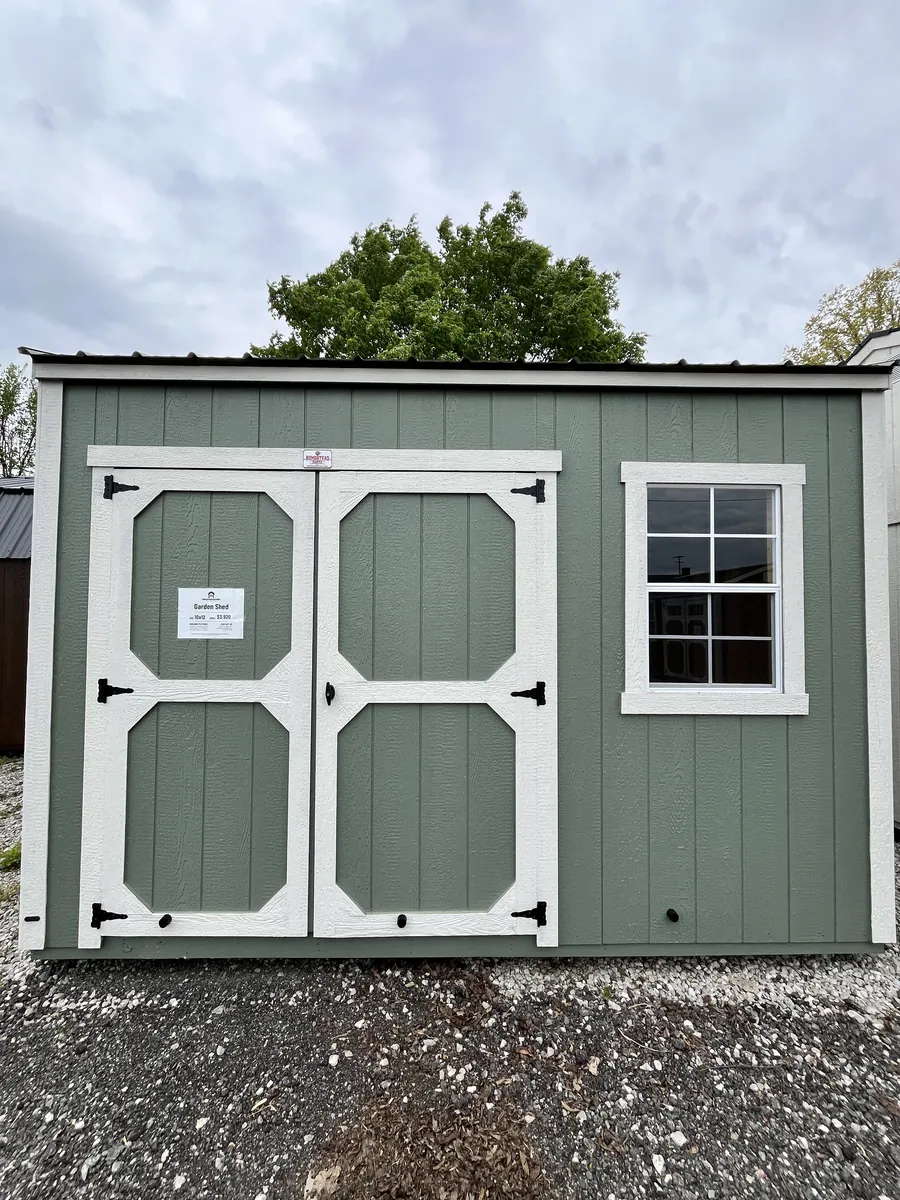 Sage green shed with white-trimmed double doors and a window on a gravel lot.