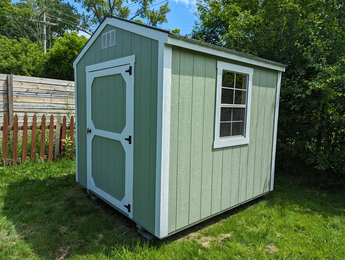 Light green utility shed with white trim and a single door in a fenced backyard.