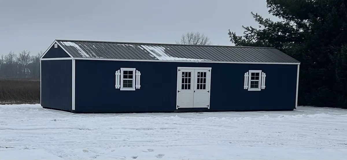 Navy blue barn-style shed with white double doors and shuttered windows in a snowy field.