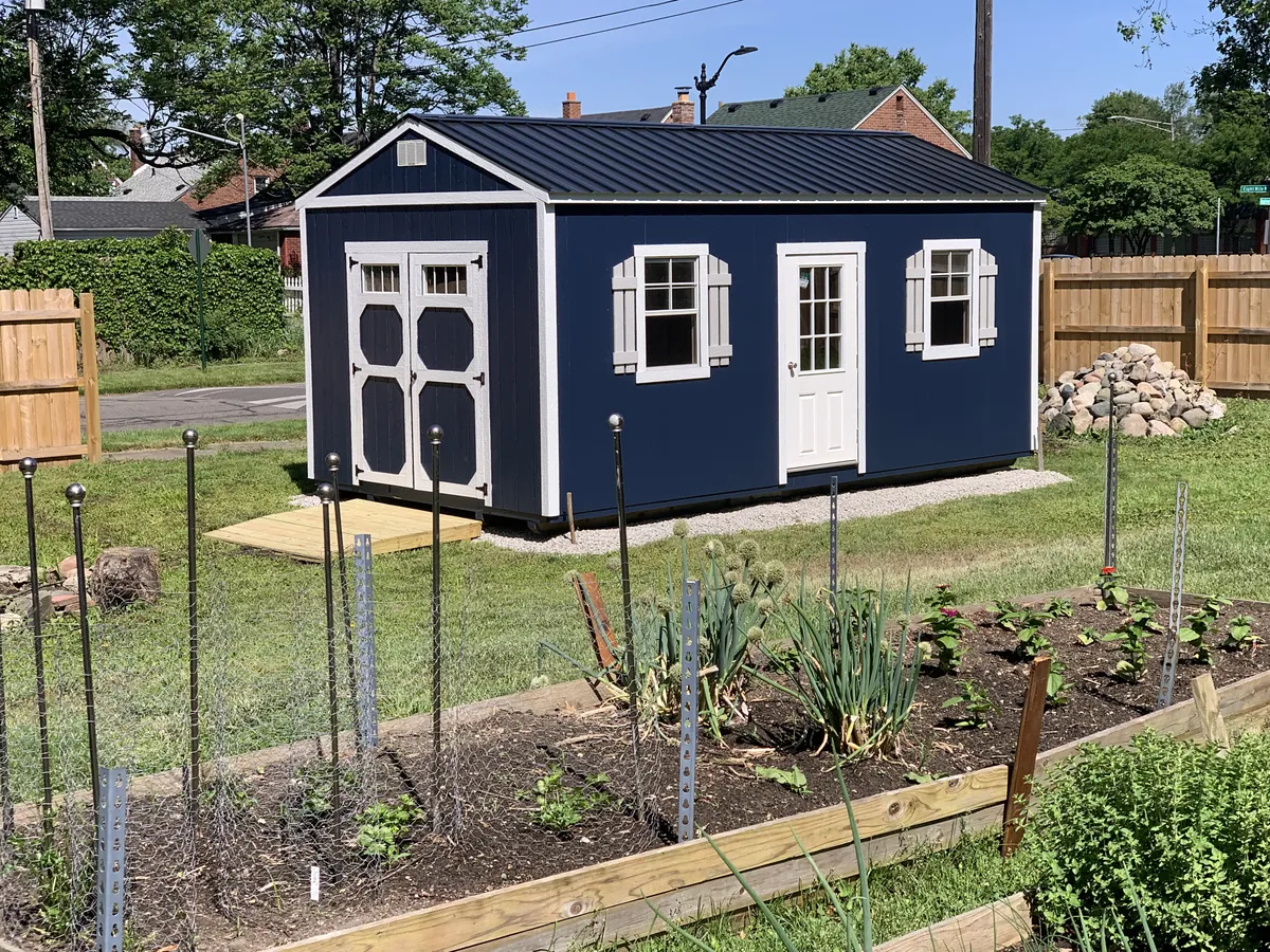 Navy shed with a white door and shutters next to a fenced vegetable garden.