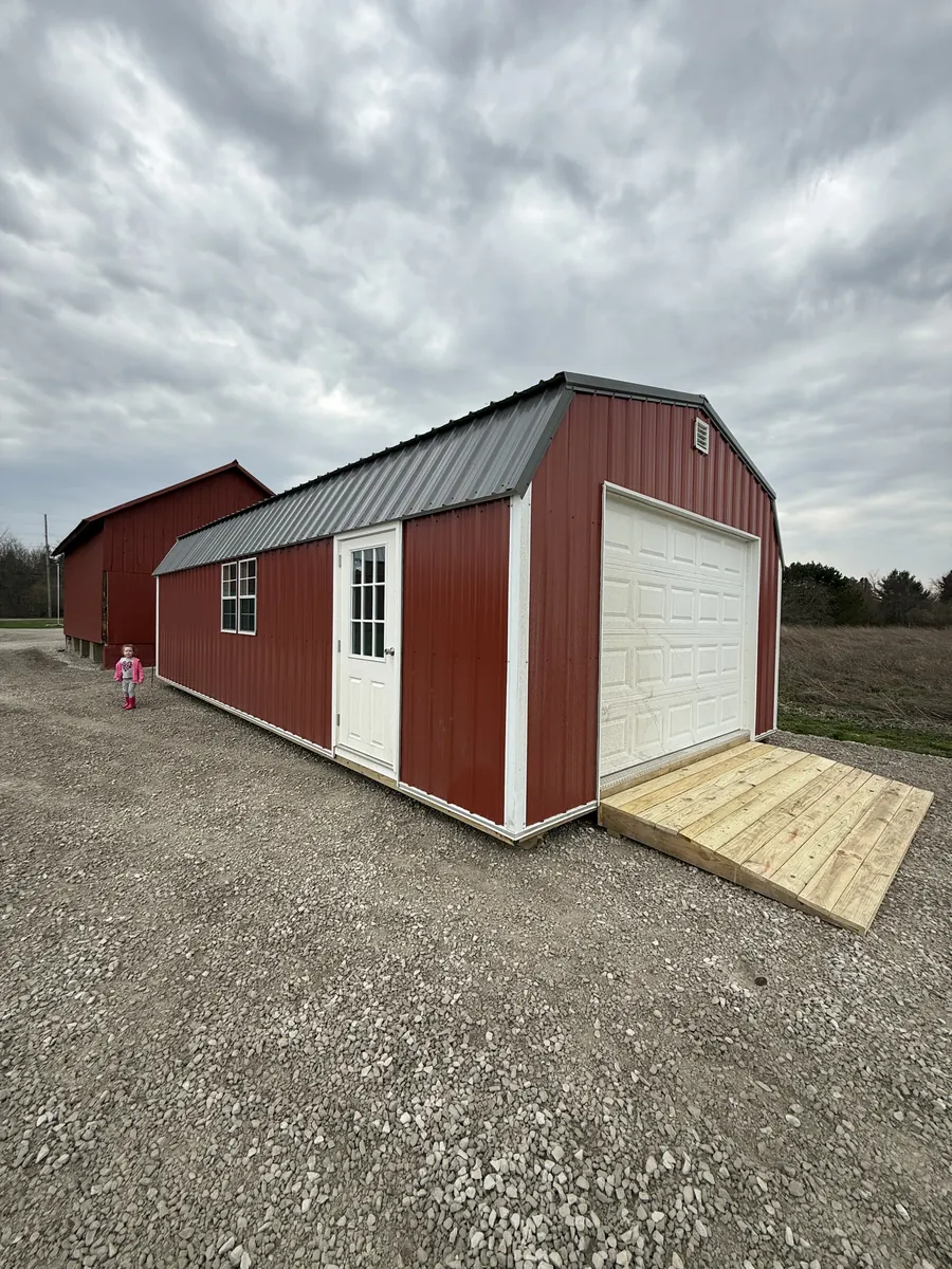 Red lofted barn garage with a white garage door, entry door, and new wooden ramp on a gravel lot.