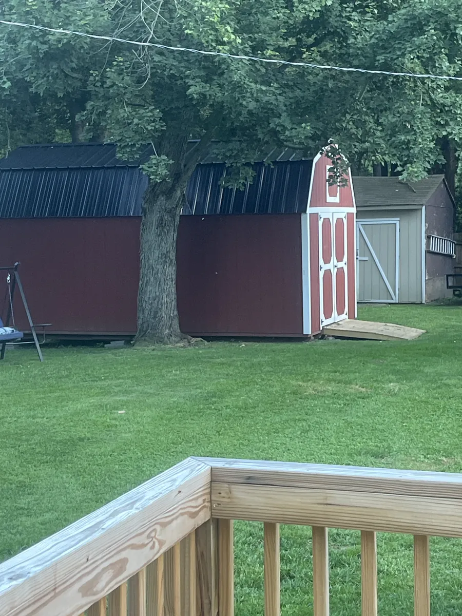 Red barn shed with white trim and a black roof on a backyard lawn, viewed from a deck.