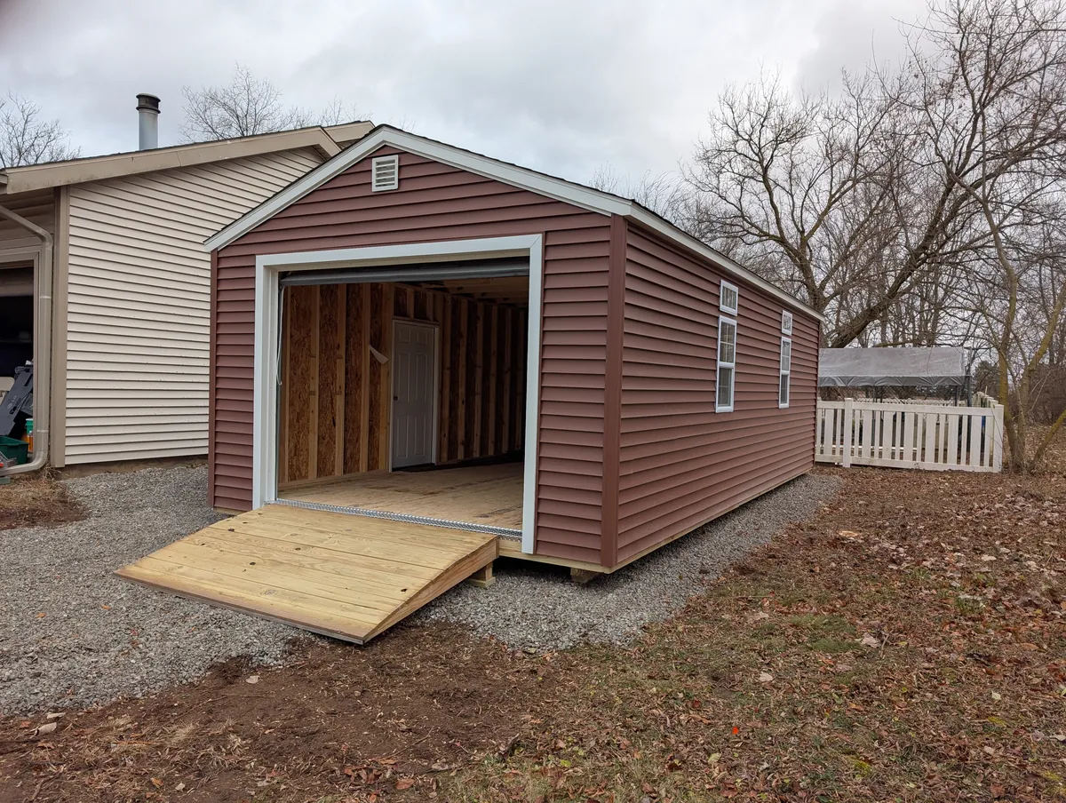 Red vinyl-sided garage shed with open garage door and wooden ramp on a gravel pad.