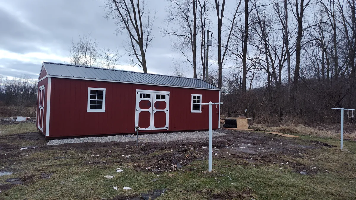 Long red garden shed with white trim and a metal roof on a muddy rural site in winter.