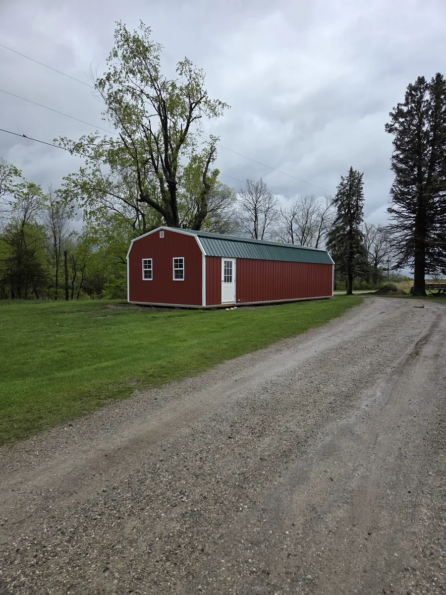 Red lofted barn shed with a green metal roof on a lawn beside a gravel driveway.