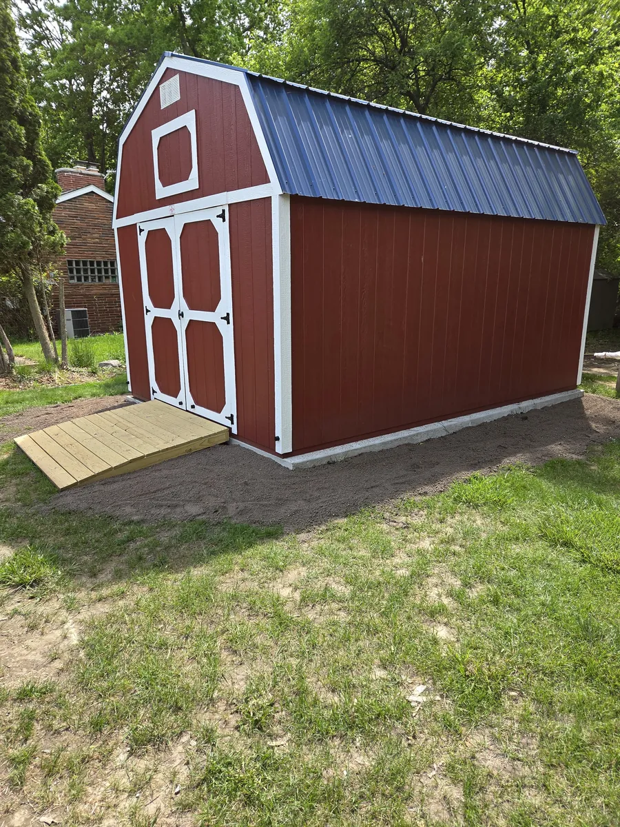 Red lofted barn shed with white trim, a metal roof, and a wooden ramp on bare ground.