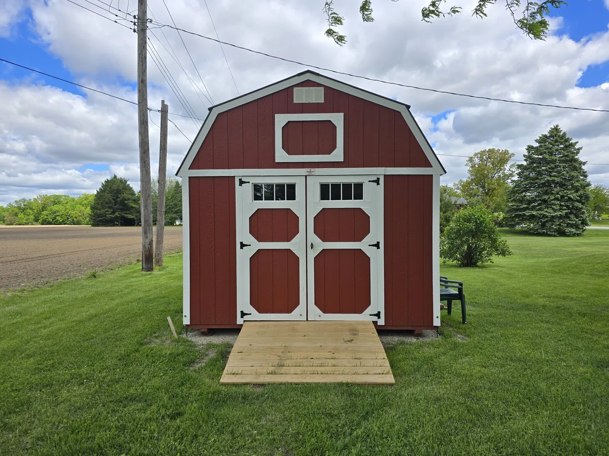 Red lofted barn shed with white-trimmed double doors and a wooden ramp in a rural yard.