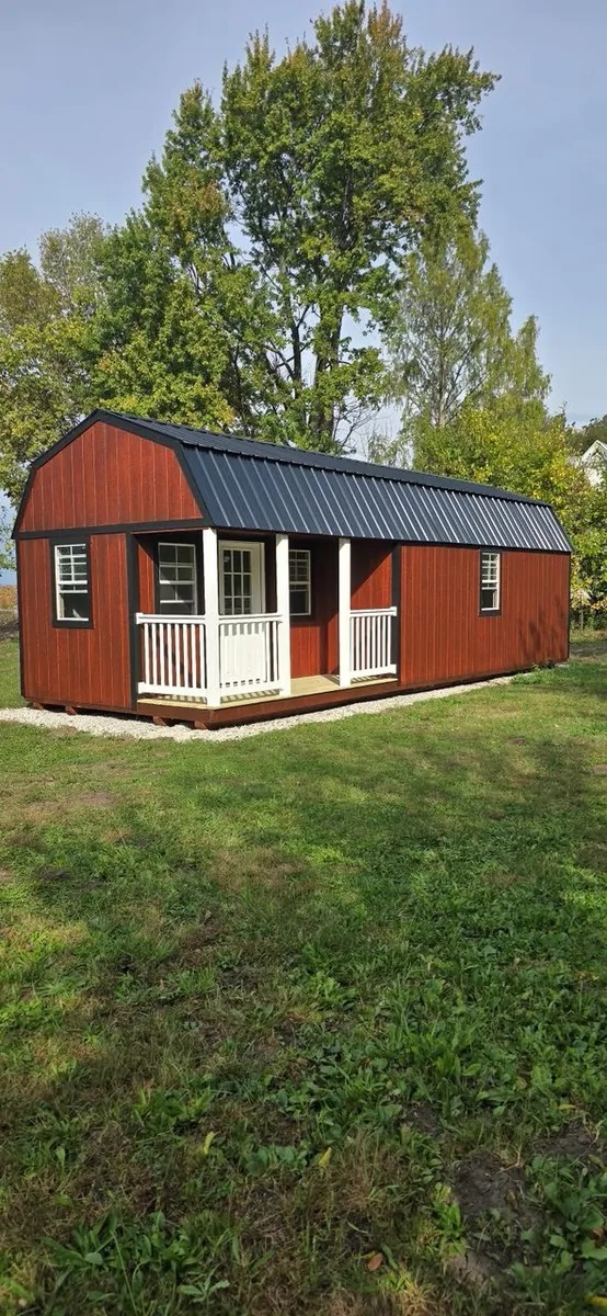 Red lofted barn cabin with a black metal roof and white porch railings on a grassy lot.
