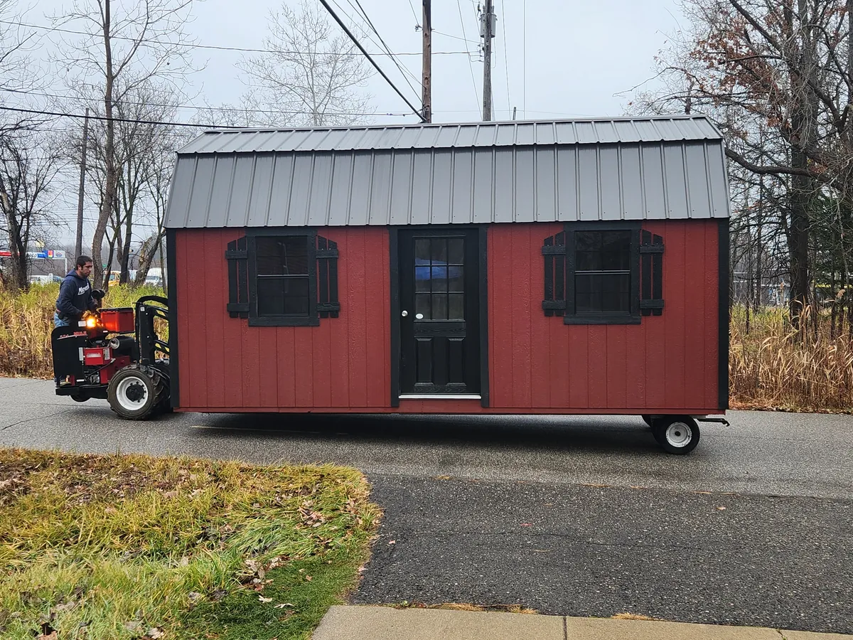 Red lofted barn shed with black shutters on wheels being moved down a street with a shed mover.