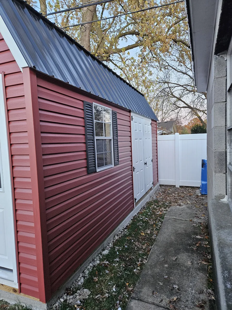Red vinyl-sided shed with a black metal roof, white door, and black shutters in a narrow side yard.