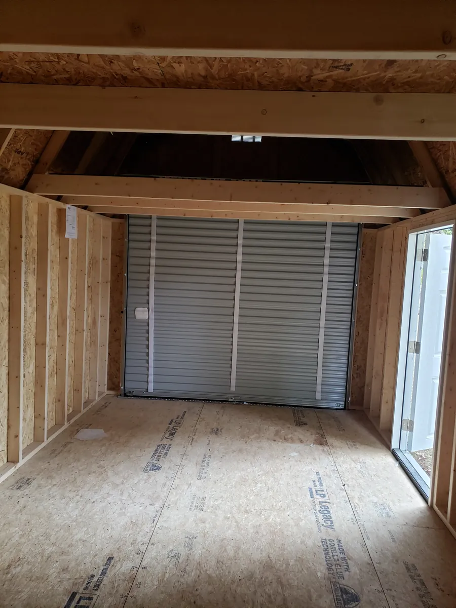Unfinished shed interior with exposed framing, a plywood floor, and a roll-up garage door.