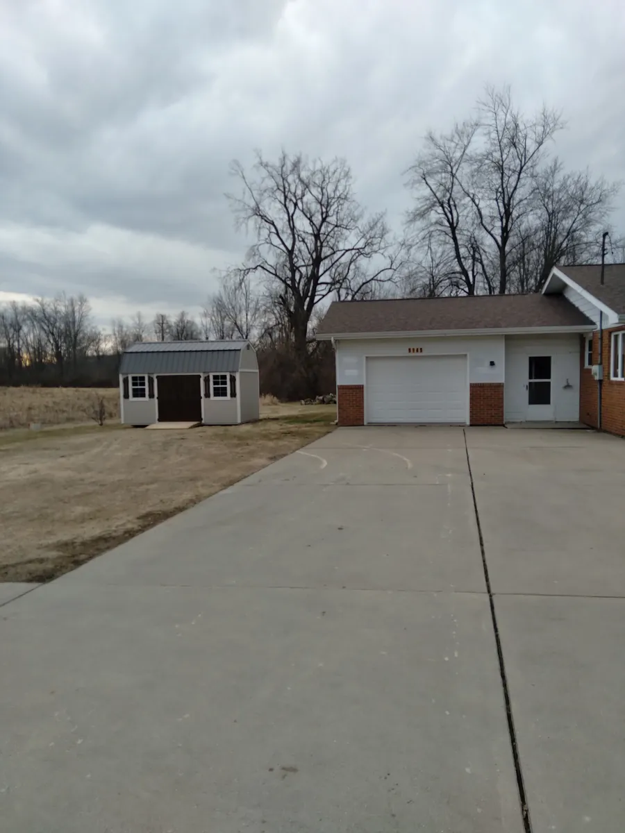Tan barn-style shed in a side yard next to a home with an attached garage and concrete driveway.