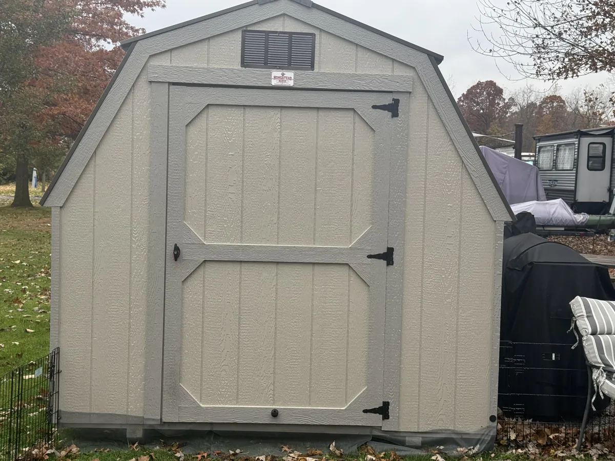 Tan barn-style shed with a single trimmed door, viewed from the front in a yard.