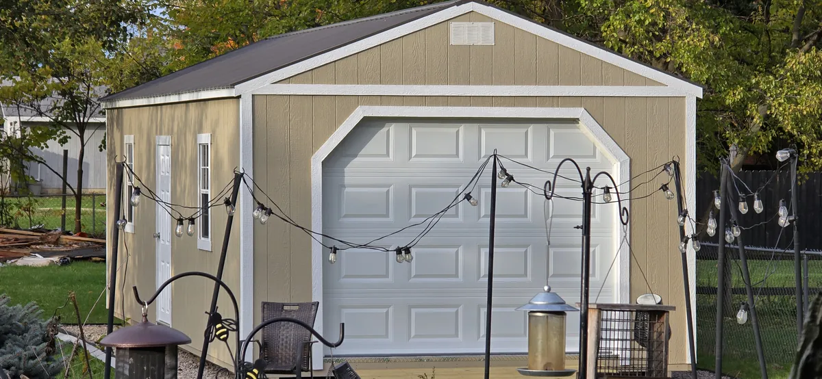 Tan garage shed with a white roll-up door and string lights on a backyard patio.